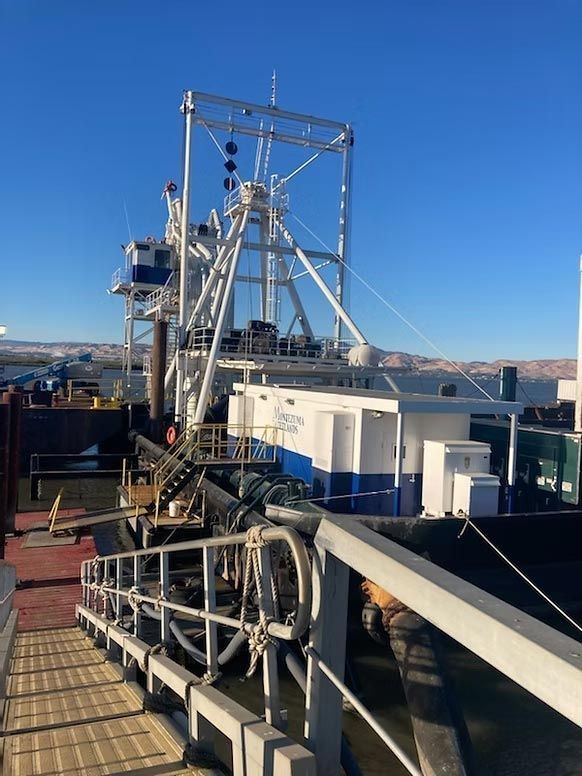 Industrial ship deck with white equipment, metal railings, and a blue sky over water and distant hills
