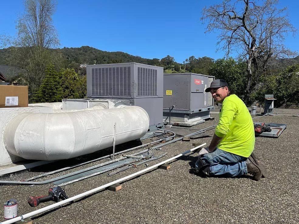 Worker kneels beside rooftop HVAC equipment and white tanks on a gravel surface under a blue sky