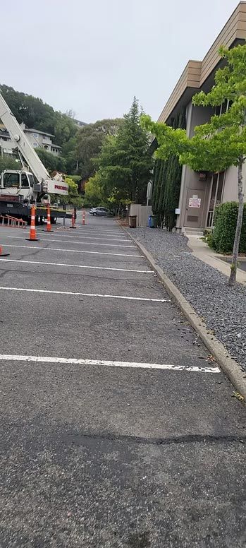 Parking lot beside a building, with orange cones, a white crane truck, and green trees lining the side