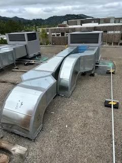 Rooftop HVAC ductwork and metal vents on a gravel surface under cloudy skies.