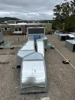 Rooftop HVAC ductwork and vents on a flat gravel roof beside a low building and trees