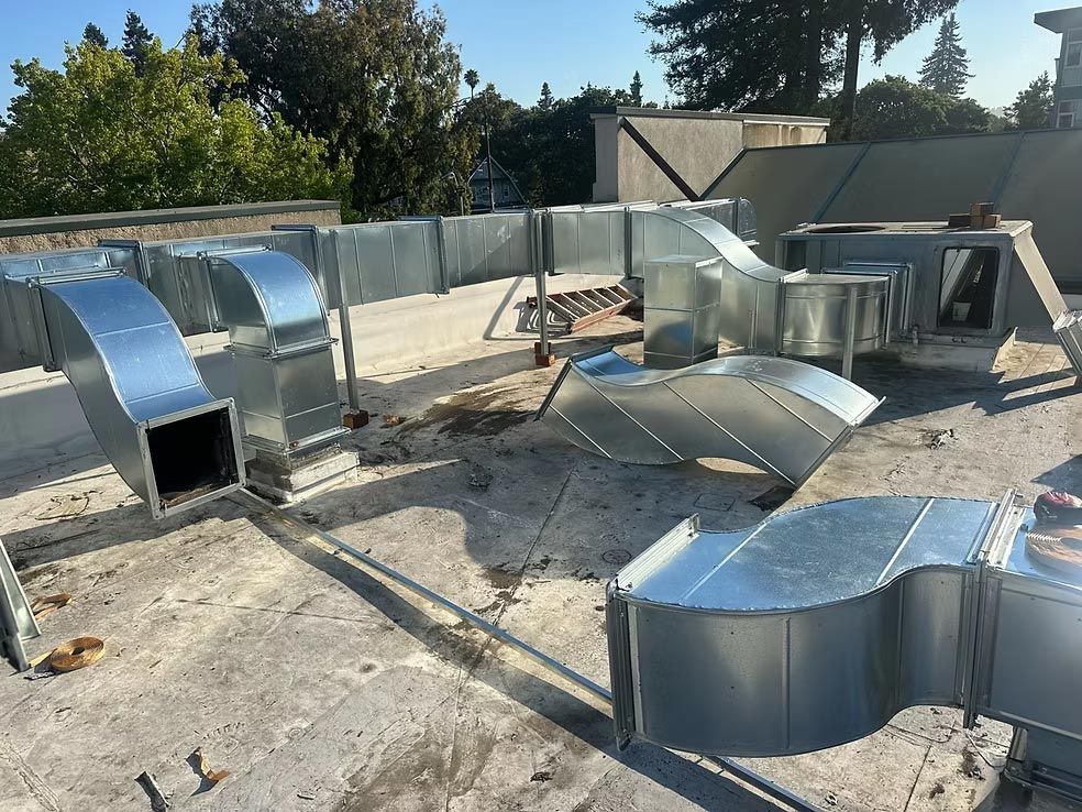 Metal HVAC duct sections scattered on a rooftop under bright sunlight, with trees and buildings in the background.