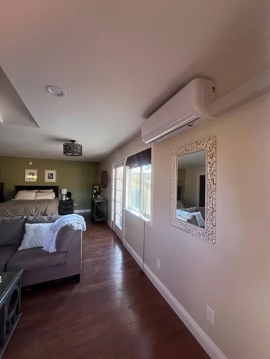 Bright bedroom with a bed, wall mirror, window, and wall-mounted air conditioner on polished wood floors