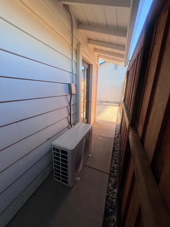 Narrow outdoor side yard with a white exterior wall, air conditioner unit, and wooden fence in bright sunlight