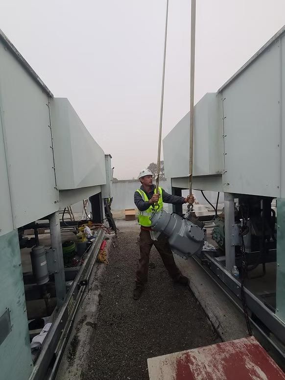 Worker in safety vest standing between large rooftop HVAC units on a gravel path.