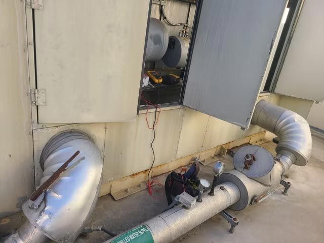 Large metal HVAC ducts and pipes on a beige rooftop wall with open equipment panels.