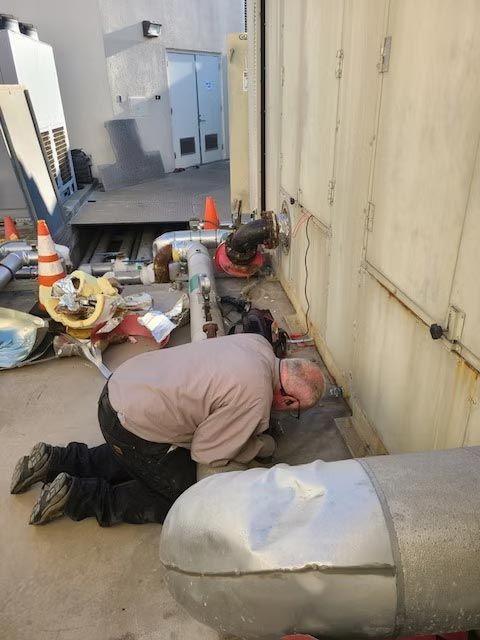 Worker crouched beside equipment in a narrow industrial alley, surrounded by tools and orange cones.