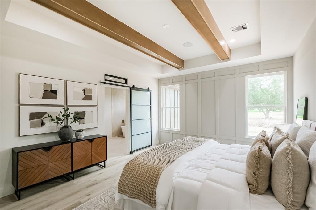 Bright bedroom with white bedding, wood-beam ceiling, dresser, and large windows with blinds