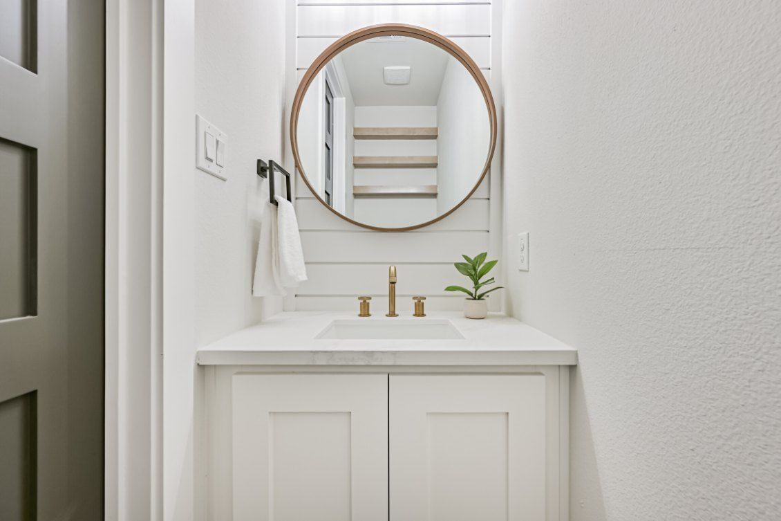 A modern white vanity with a marble countertop, gold faucet, round mirror, and a small plant on a shiplap wall.
