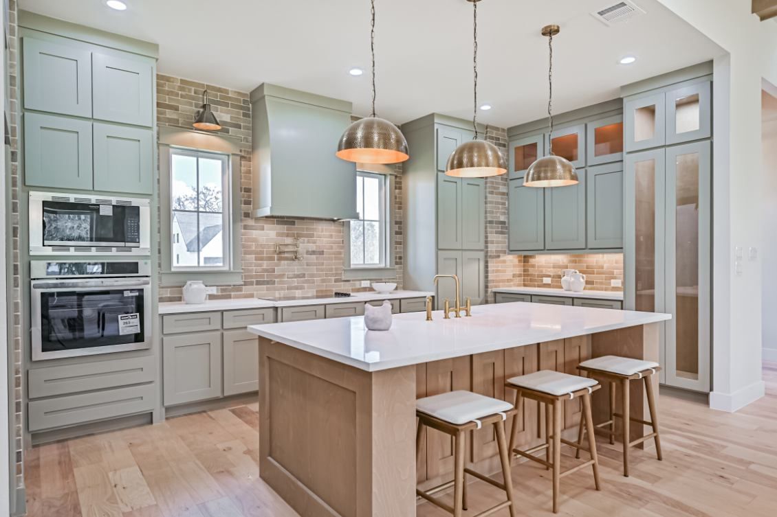 A modern kitchen featuring light blue cabinetry, stone accents, a white-topped island with three stools, and gold lighting.