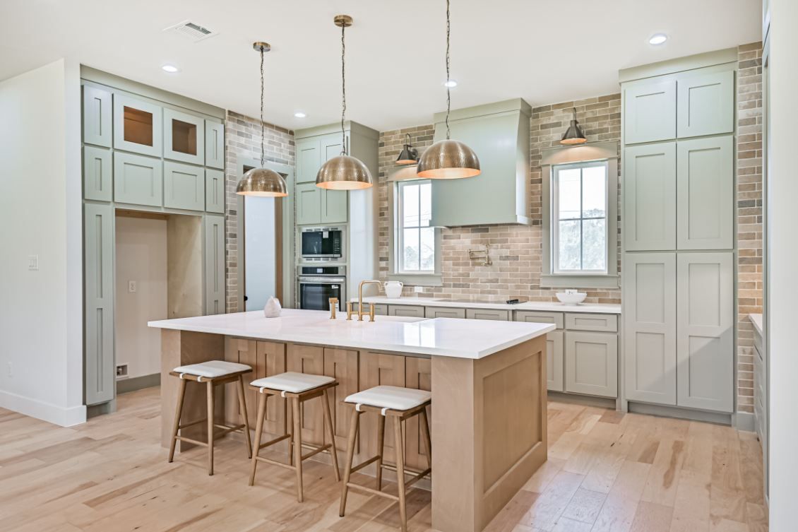 A modern kitchen with light green cabinets, a wood-toned island with three stools, and stone accent walls.