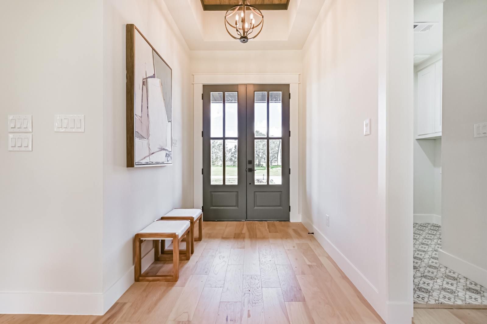A bright entryway with light wood floors, a dual-door entrance, a wall-mounted abstract art piece, and two wooden benches.