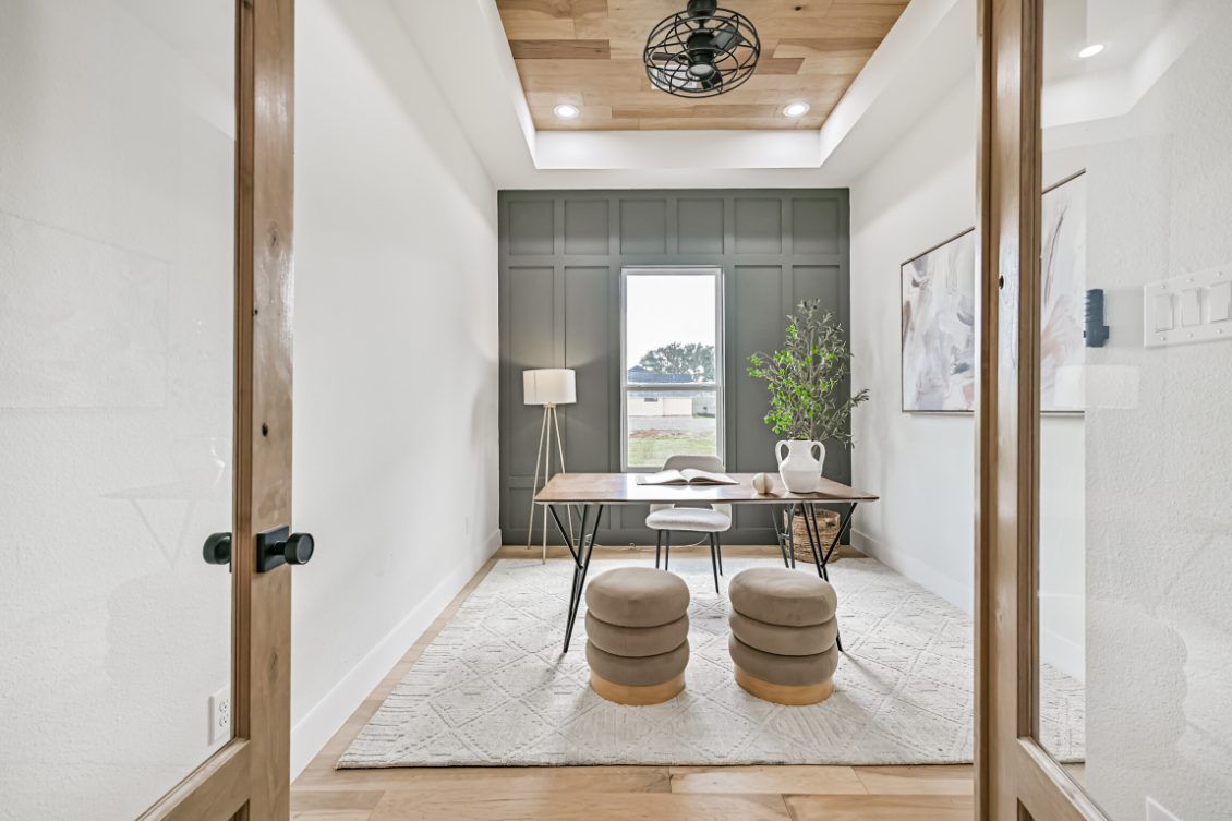 A home office with a wooden ceiling, a dark accent wall, a central desk, two stools, and a light rug, viewed through doors.