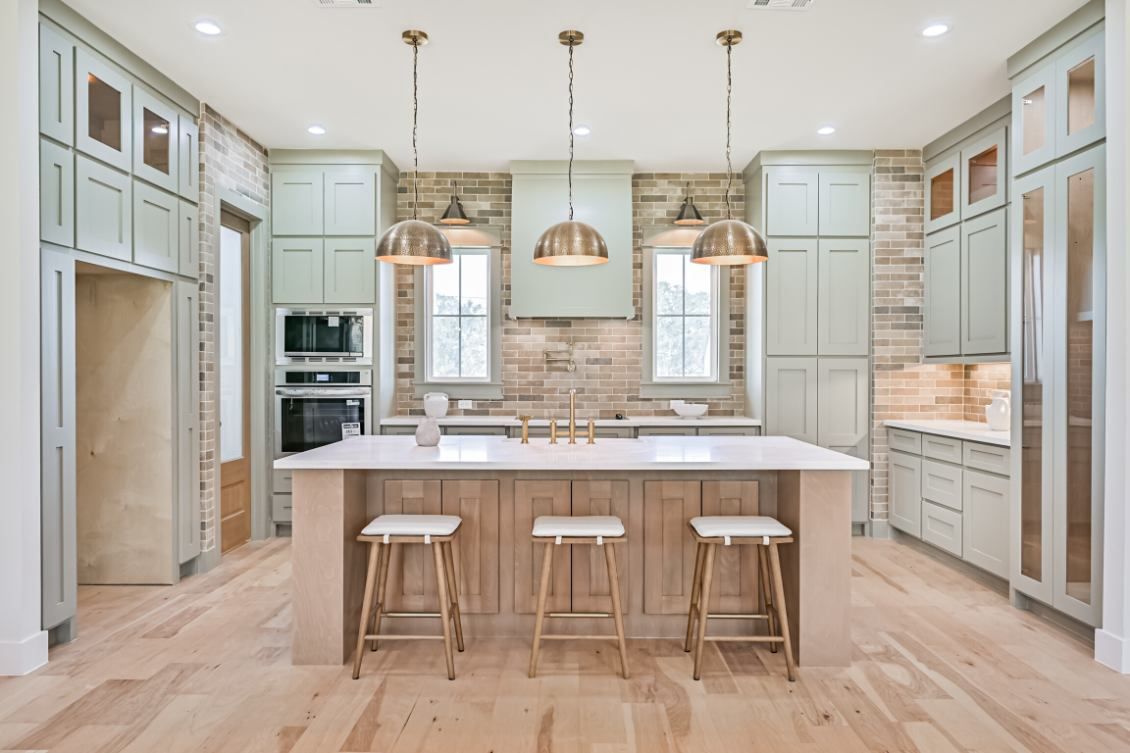 A spacious kitchen with sage green cabinets, wood-toned island with three stools, pendant lighting, and stone backsplash.
