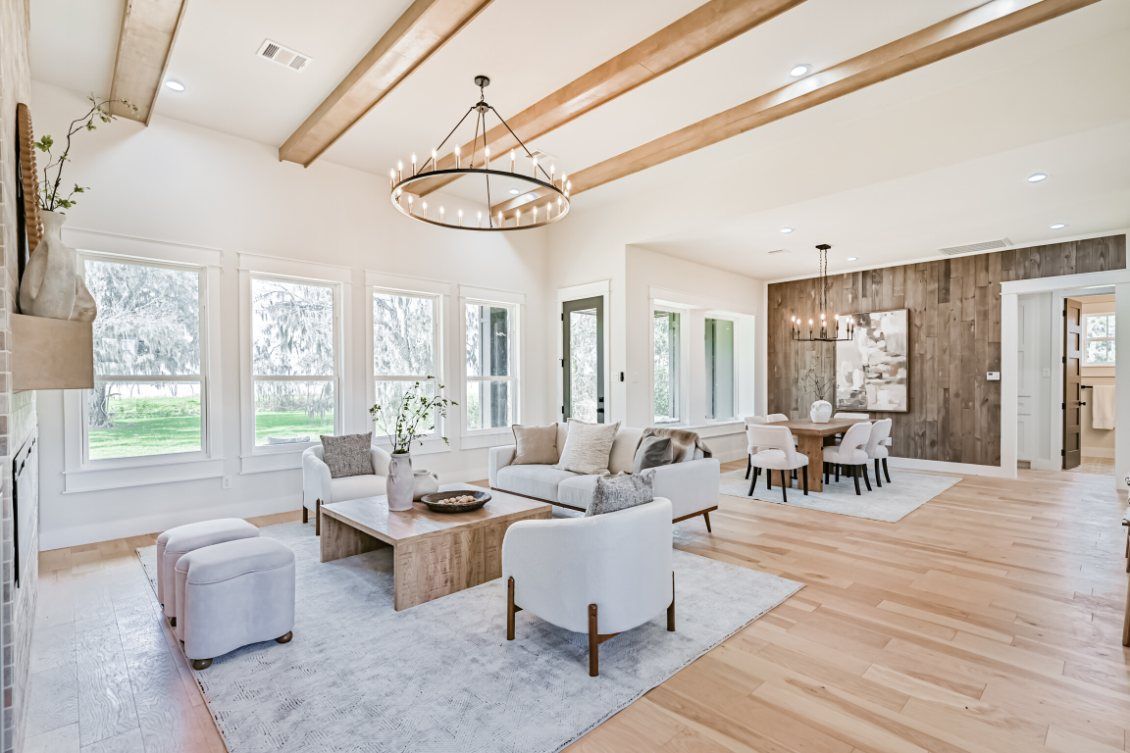 A bright, open-concept living and dining room with white furniture, wooden ceiling beams, and a farmhouse-style chandelier.