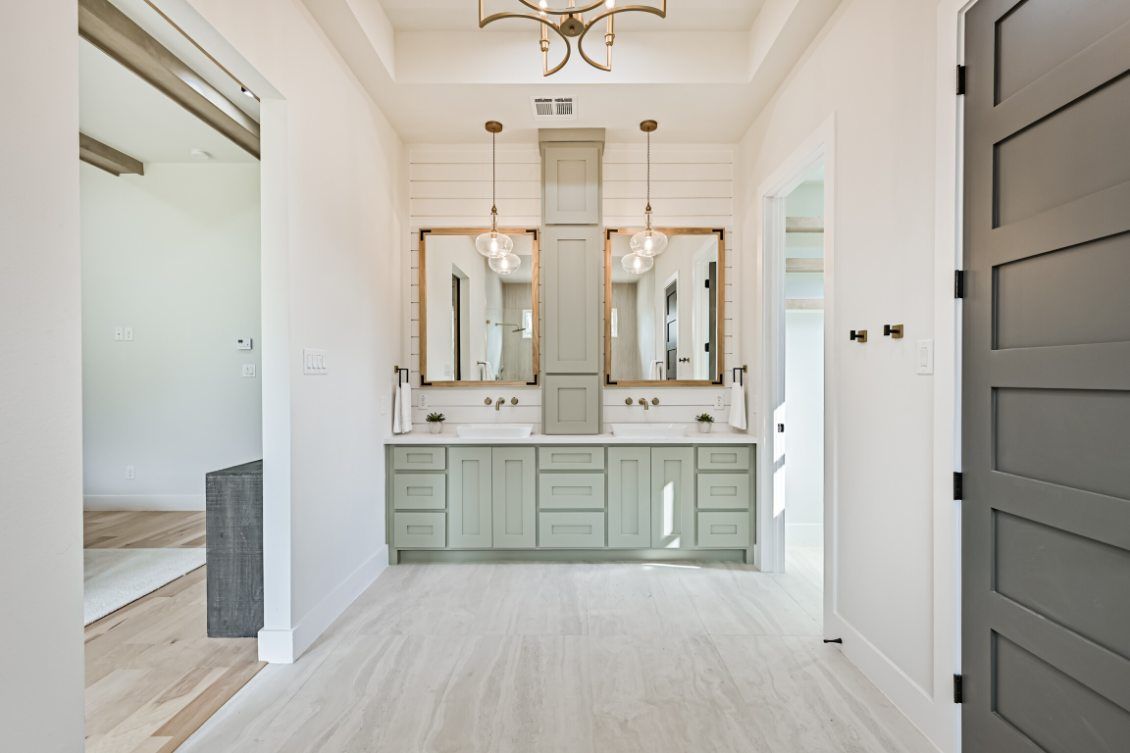 Modern bathroom with a double vanity, sage green cabinets, gold mirrors, and a light wooden floor.