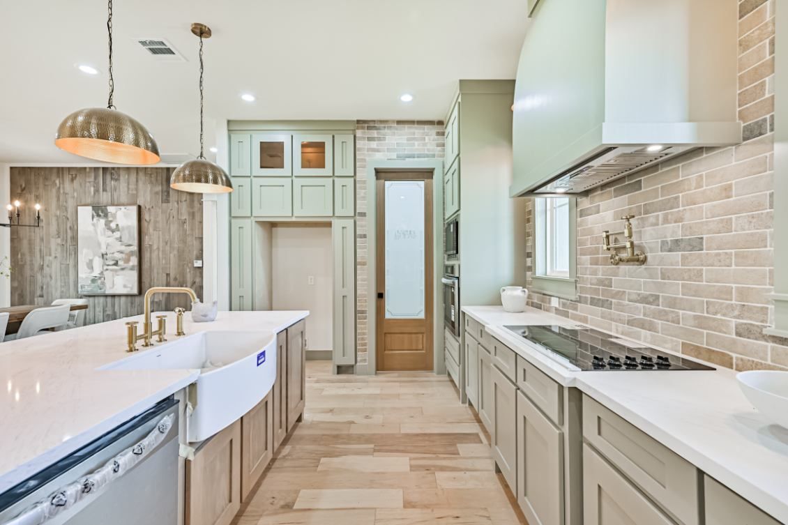 Modern kitchen with sage green cabinets, light wood flooring, brick backsplash, and a large white farmhouse sink.