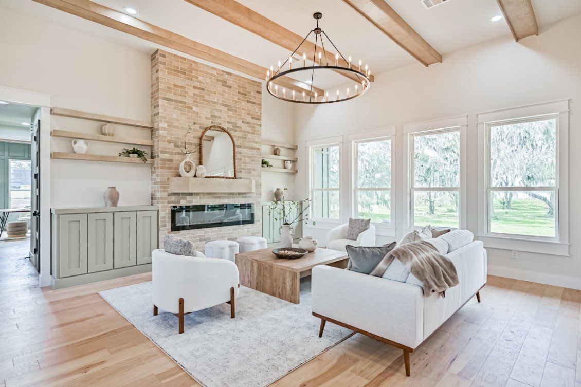A bright, modern living room featuring a stone fireplace, white sofa and armchair, wooden beams, and a round chandelier.