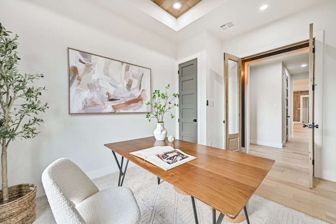 A bright home office with a wooden desk, white chair, abstract art, and an olive tree under a tray ceiling.