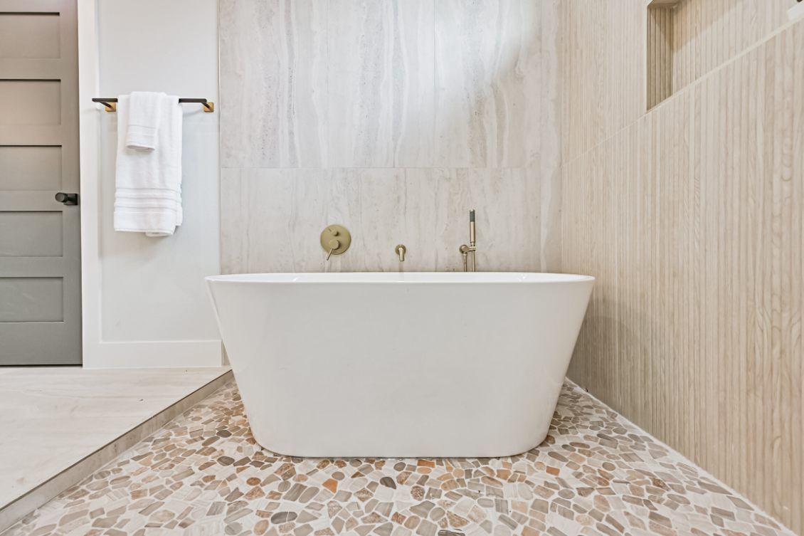 Modern bathroom with a white freestanding bathtub on a stone pebble floor, featuring beige tiled walls and brass fixtures.