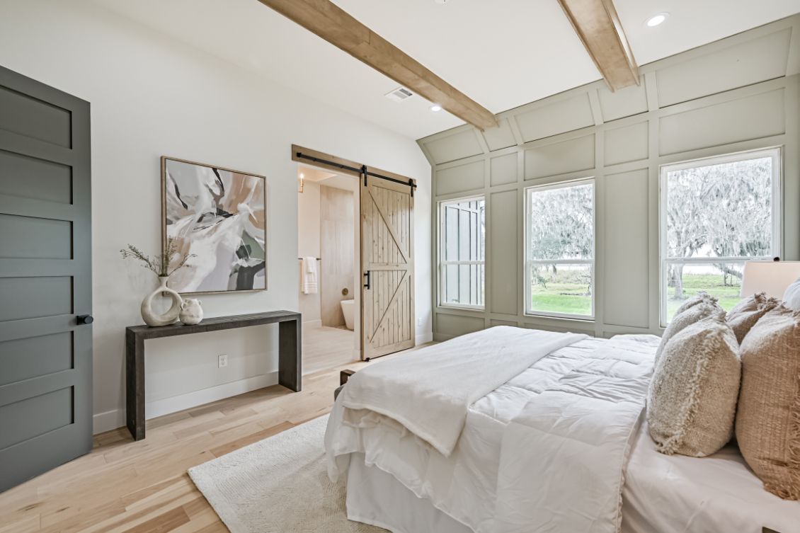A bedroom with light wood floors, a bed with cream bedding, a gray door, and a sliding wooden door leading to a bathroom.