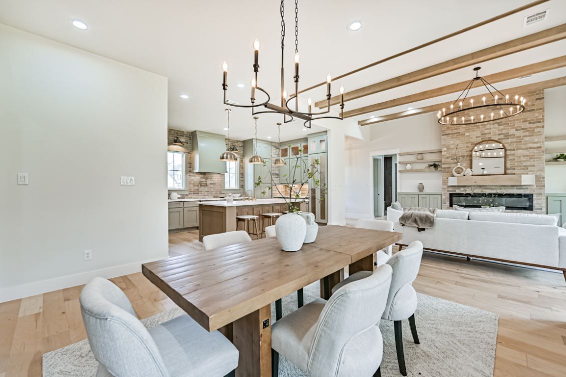 An open-concept dining area with a wood table, light gray chairs, and a chandelier, leading into a kitchen and living room.