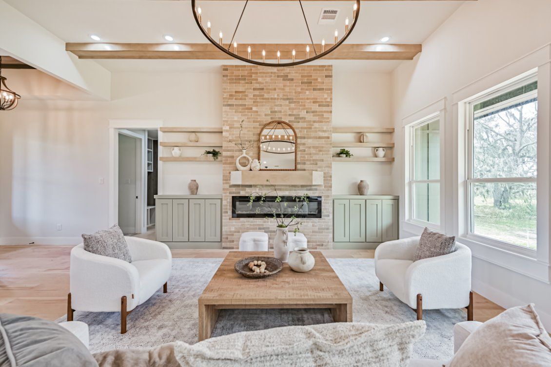 A bright living room with a stone fireplace, two white armchairs, a wooden coffee table, and a large metal chandelier.
