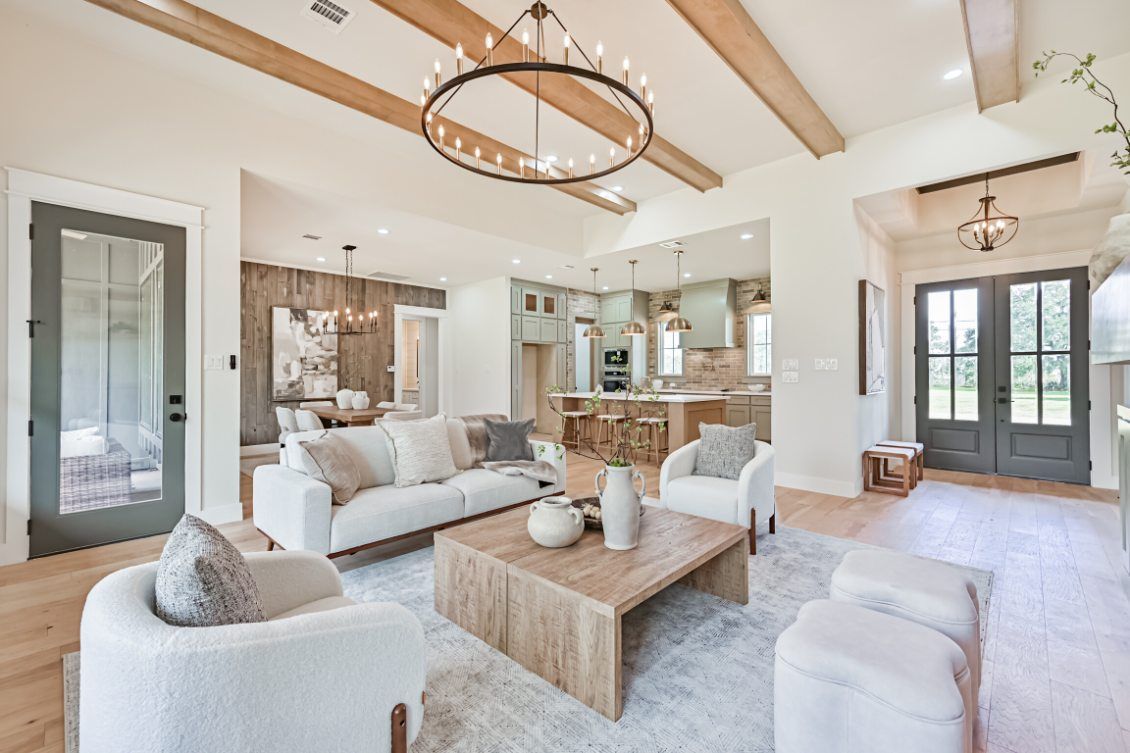 A bright living room with white sofas, a wooden coffee table, a wagon wheel chandelier, and open-plan kitchen background.