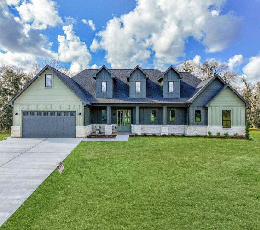 A single-story suburban house with sage green siding, dark gray roof, and a white stone base under a blue, cloudy sky.