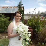 Bride in white dress holding a bouquet, standing outdoors near a rustic building and garden.