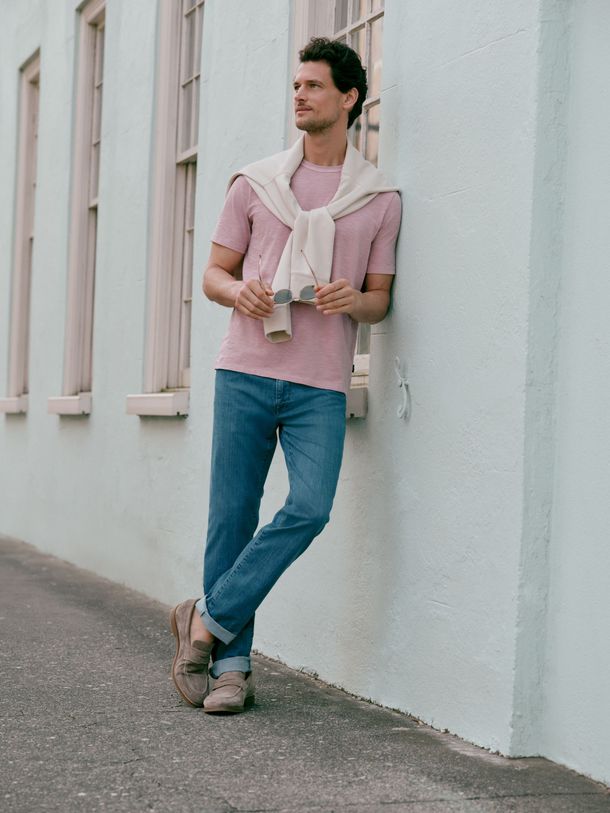 Man leaning against a white wall outdoors, wearing a pink shirt, blue jeans, and holding a drink.