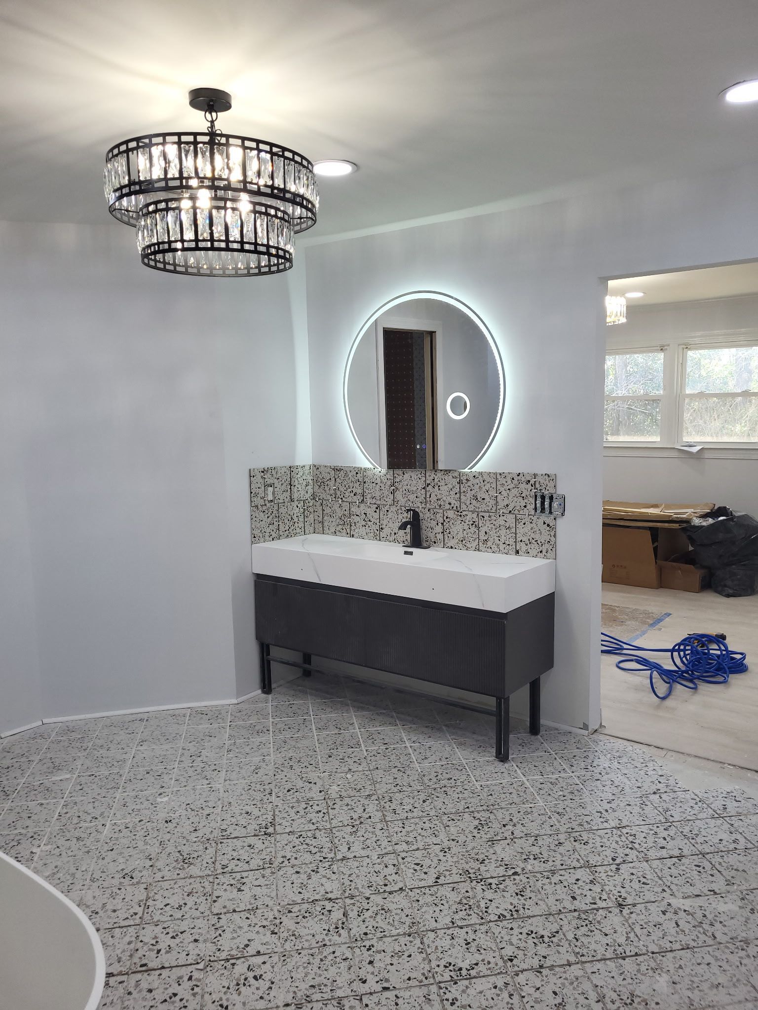 Modern bathroom with terrazzo floor, round backlit mirror, black vanity, and chandelier