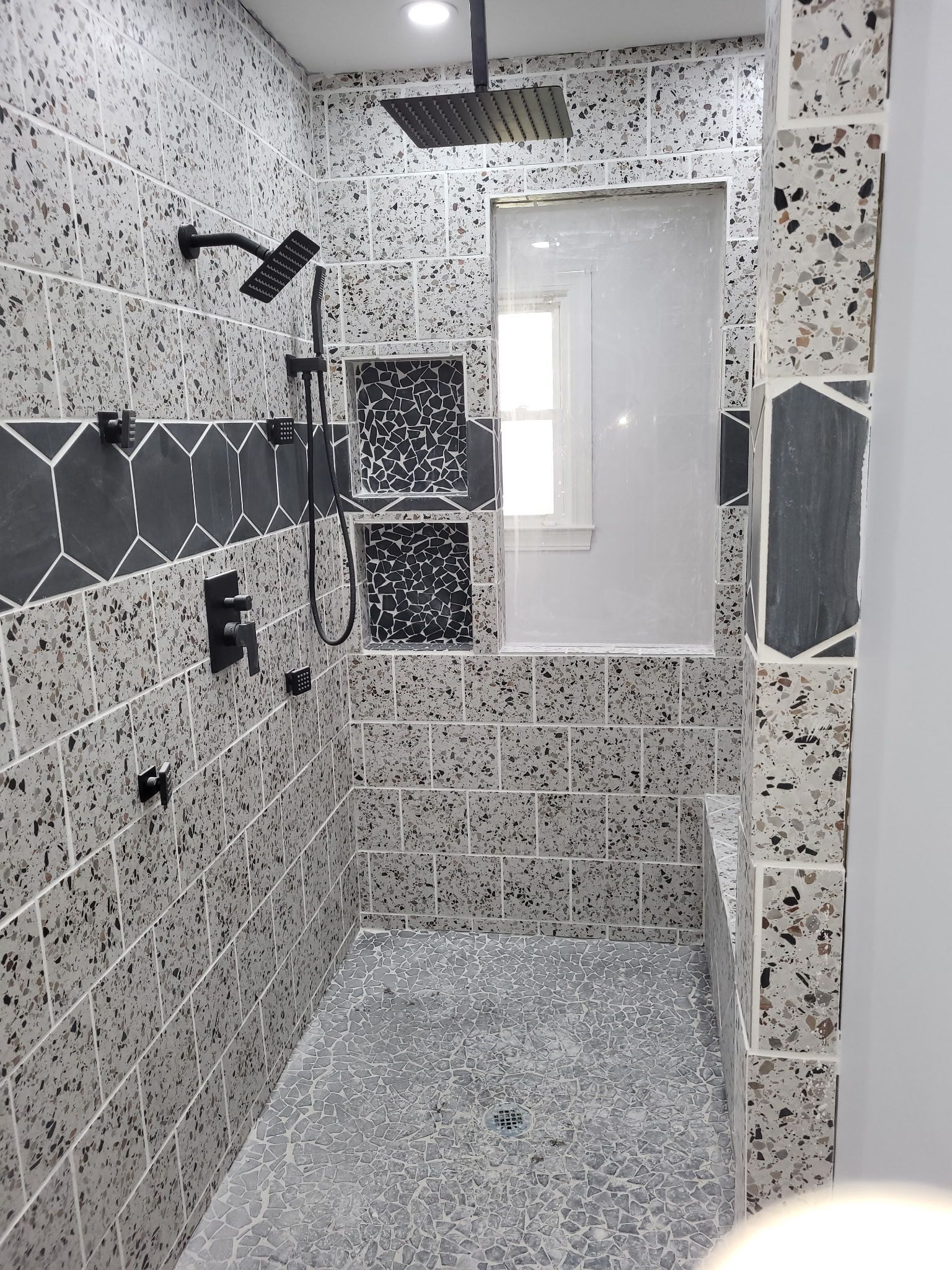 Small tiled shower room with white door, gray floor, and black-and-white mosaic walls.