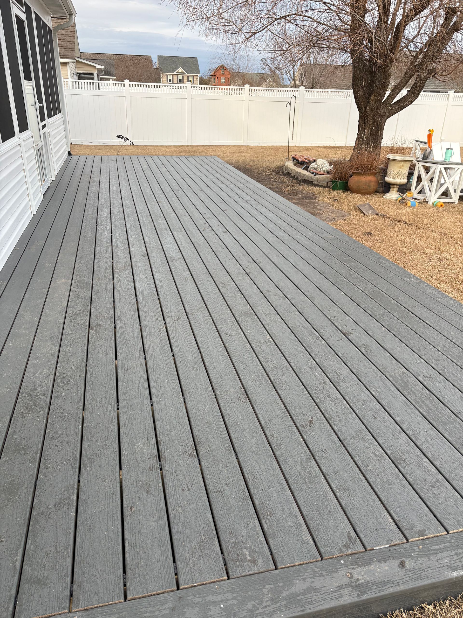 Gray wooden deck in a backyard with a white fence, bare tree, and patio chairs along the right side