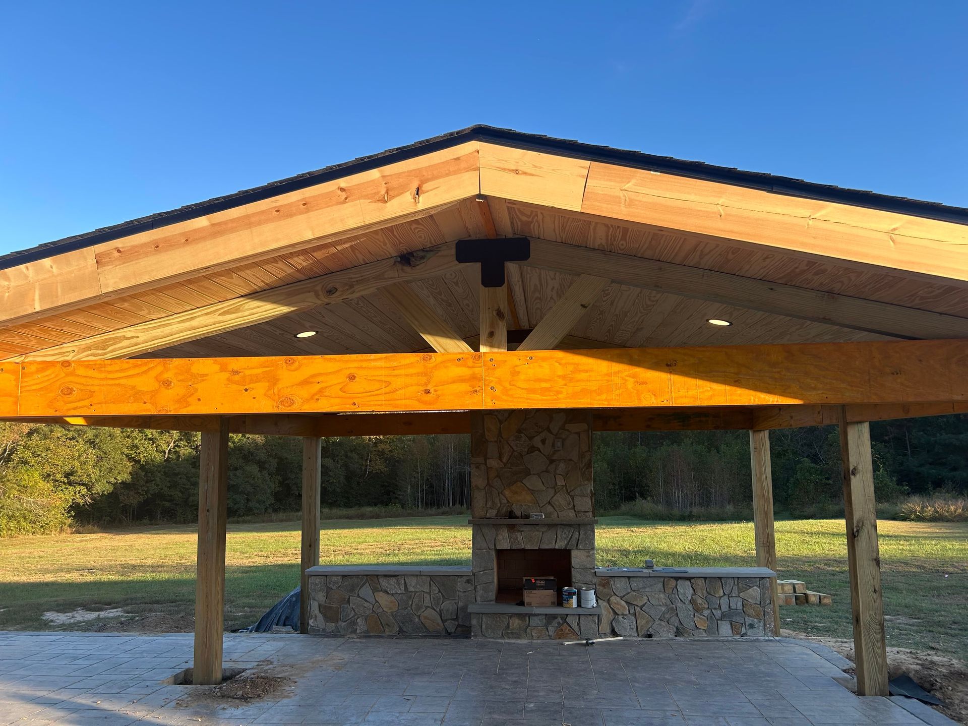 Open-air pavilion with wooden roof, stone fireplace, and supporting posts in a grassy field