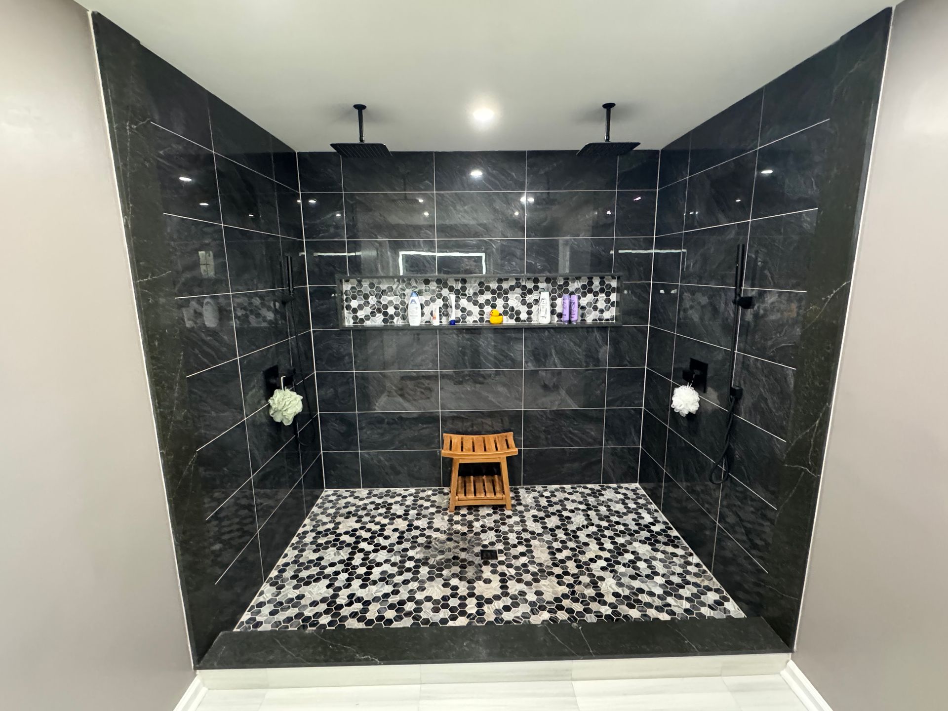 Black-tiled shower stall with mosaic floor, bench, and two showerheads under a recessed ceiling.