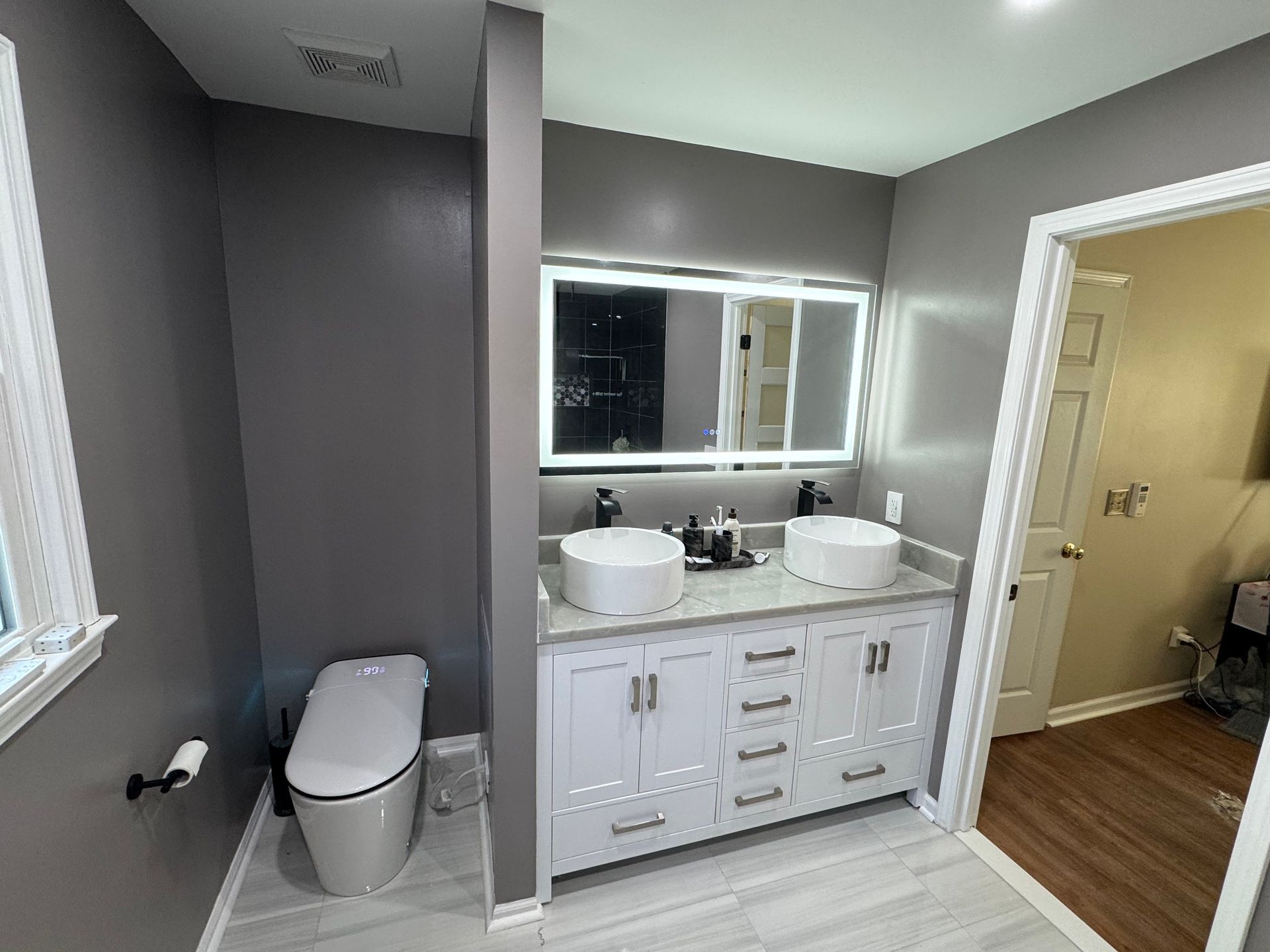 Modern bathroom with white vanity, dual sinks, mirror, toilet, and open doorway to another room