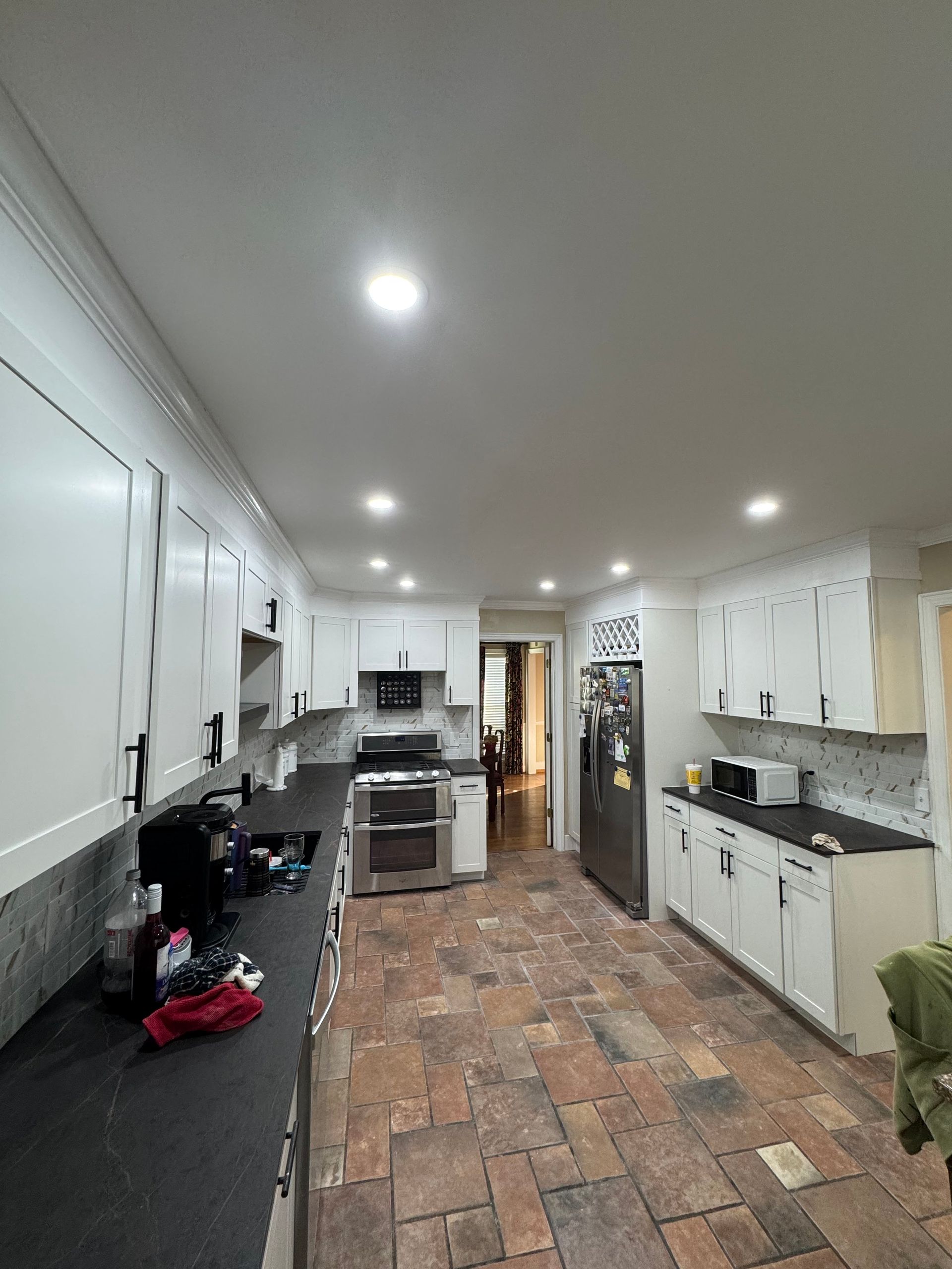 Bright kitchen with white cabinets, black counters, brick floor, and recessed ceiling lights.