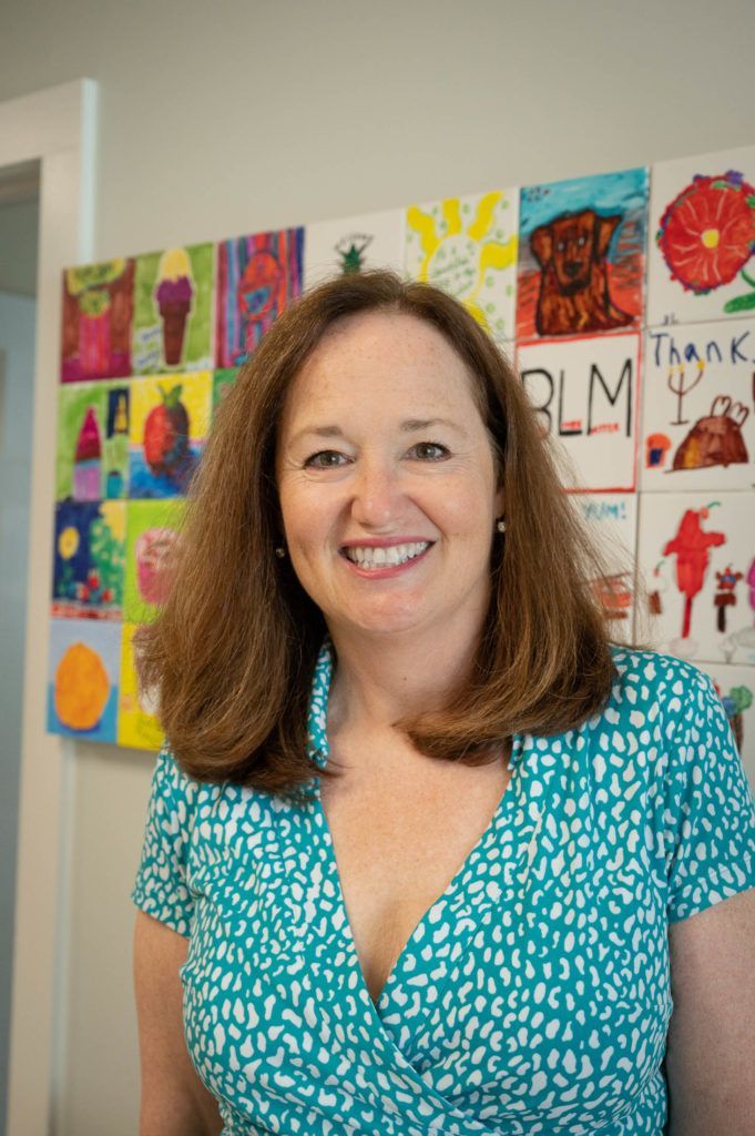 A woman in a blue shirt is smiling in front of a wall of paintings.