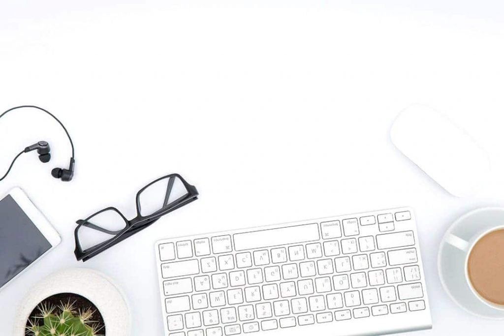 A white desk with a keyboard , glasses , headphones , a cup of coffee and a potted plant.