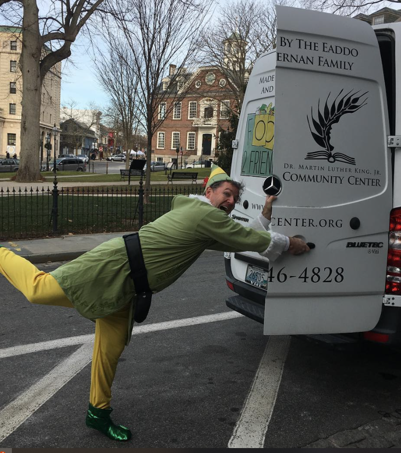 A man dressed as an elf is standing next to a van that says community center