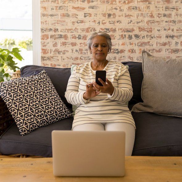 A woman is sitting on a couch using a laptop and a cell phone.