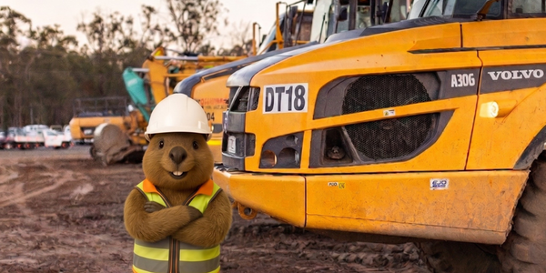 Construction mascot in hard hat stands before a yellow Volvo excavator at a muddy worksite