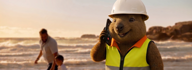 Person in hard hat and safety vest speaking on a phone at a beach during sunset, with another person in the background.