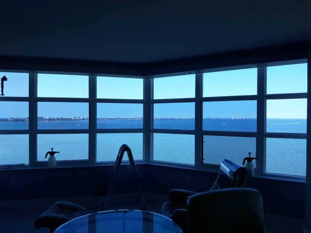 Blue-tinted view through large bay windows over a waterfront at dusk, with dark silhouettes of chairs indoors.