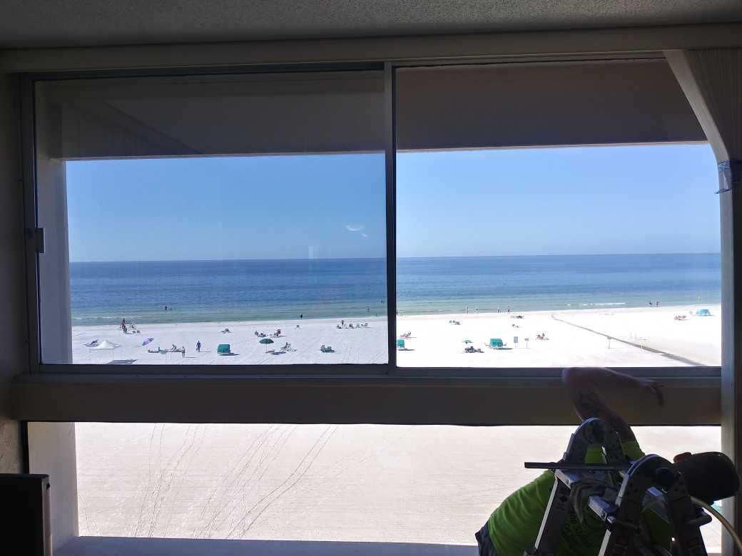 Beach and ocean seen through a window from an indoor balcony, with white sand and blue sky.