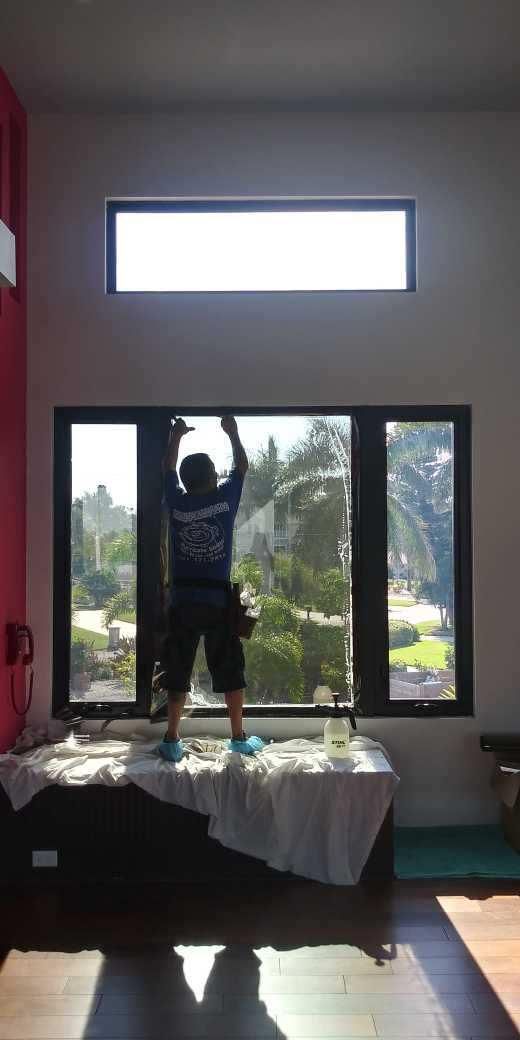 Person standing on a table in a sunlit room, arms raised in front of large windows.