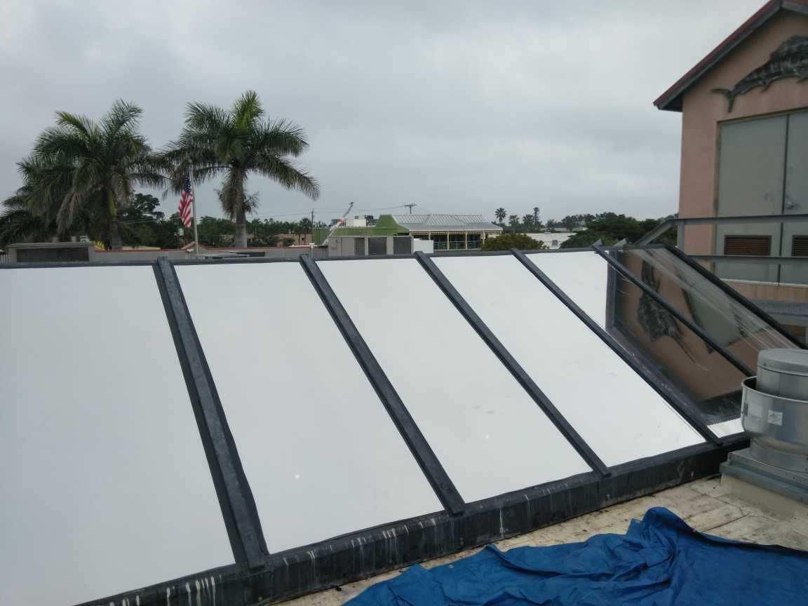 Flat rooftop solar water heater panels under an overcast sky, with palm trees in the background