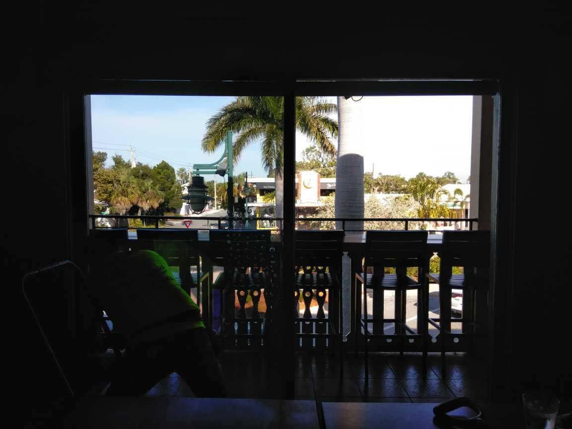Balcony view through a shaded doorway toward a sunny palm-lined pool area and white buildings