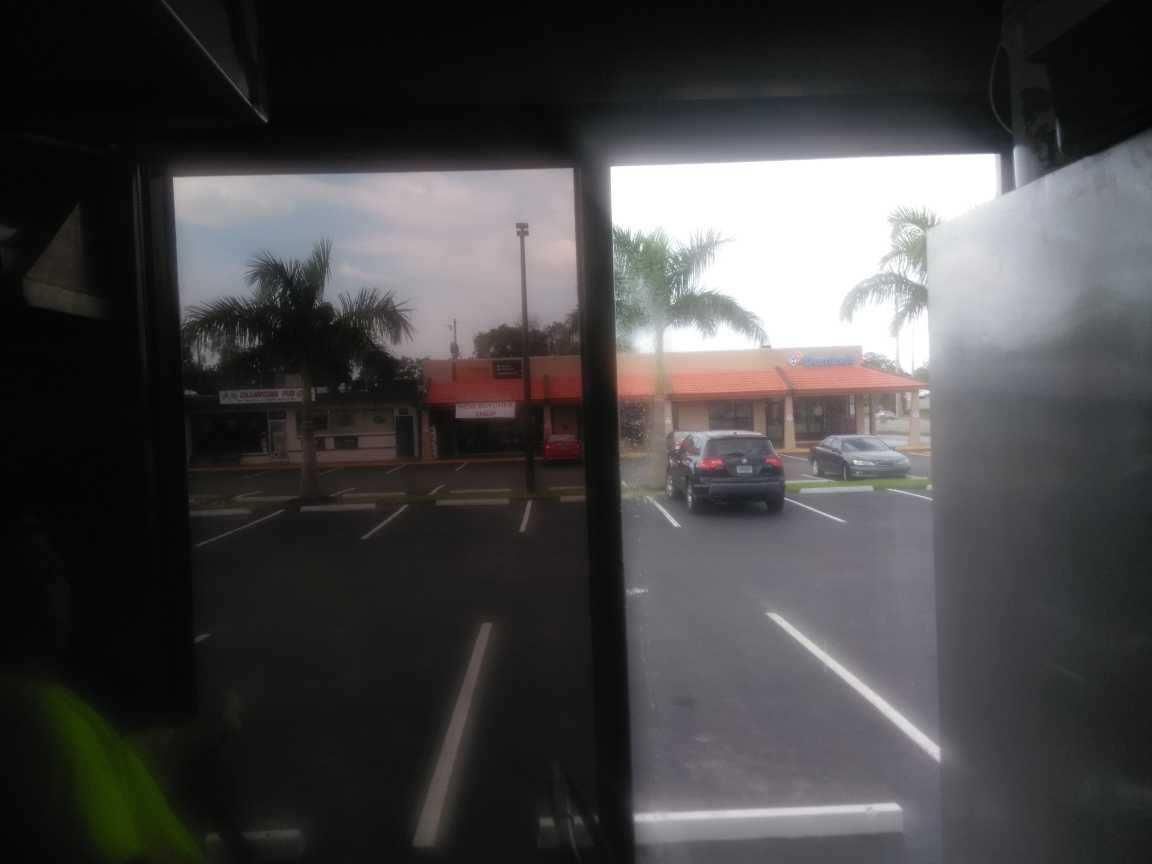View from inside a bus toward a parking lot and storefronts on a rainy day